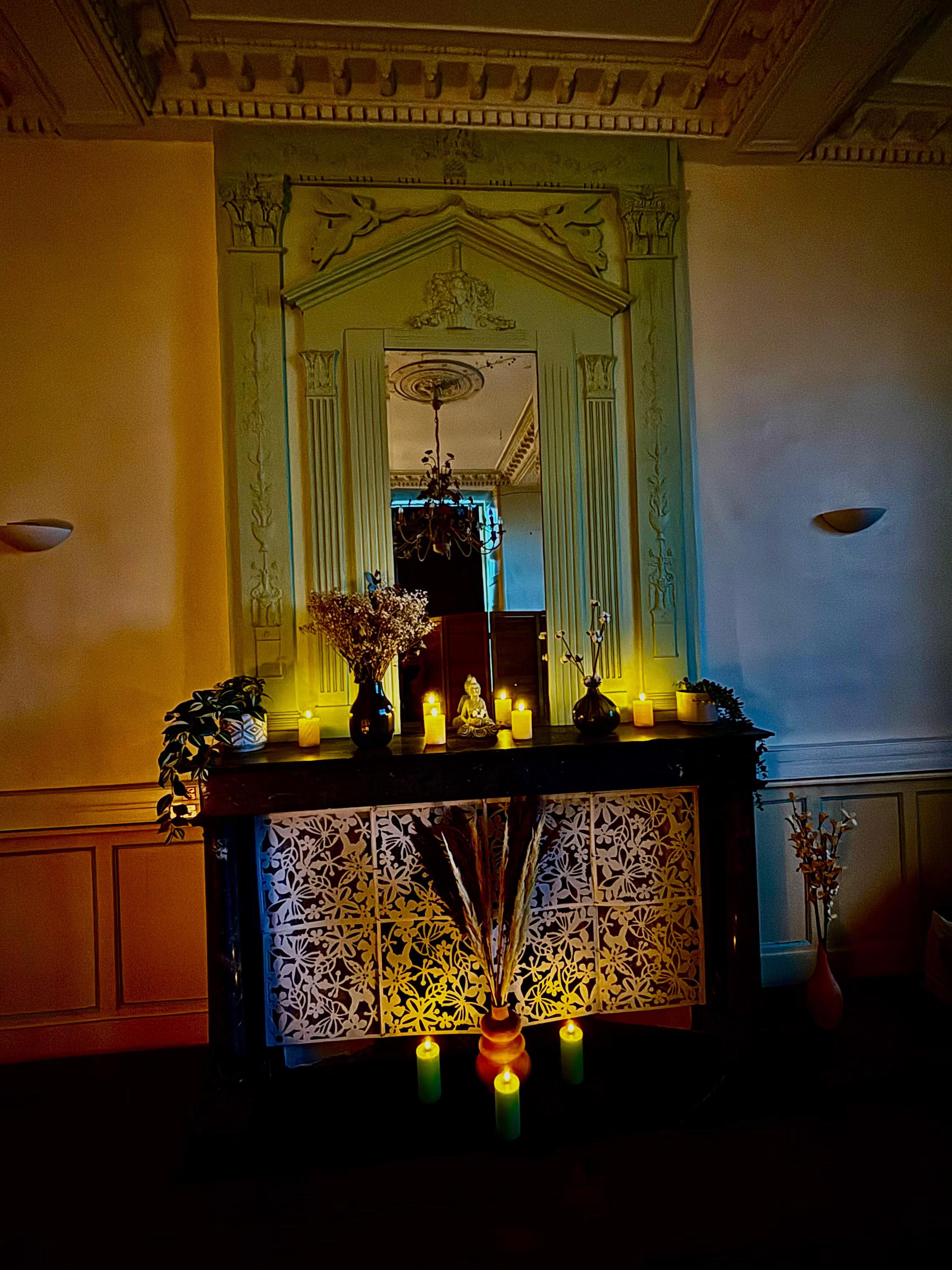 Le Logis de la Treille : hall d’accueil élégant avec un grand miroir ornementé, une cheminée décorative, des bougies allumées et des compositions florales dans une ambiance chaleureuse et apaisante.