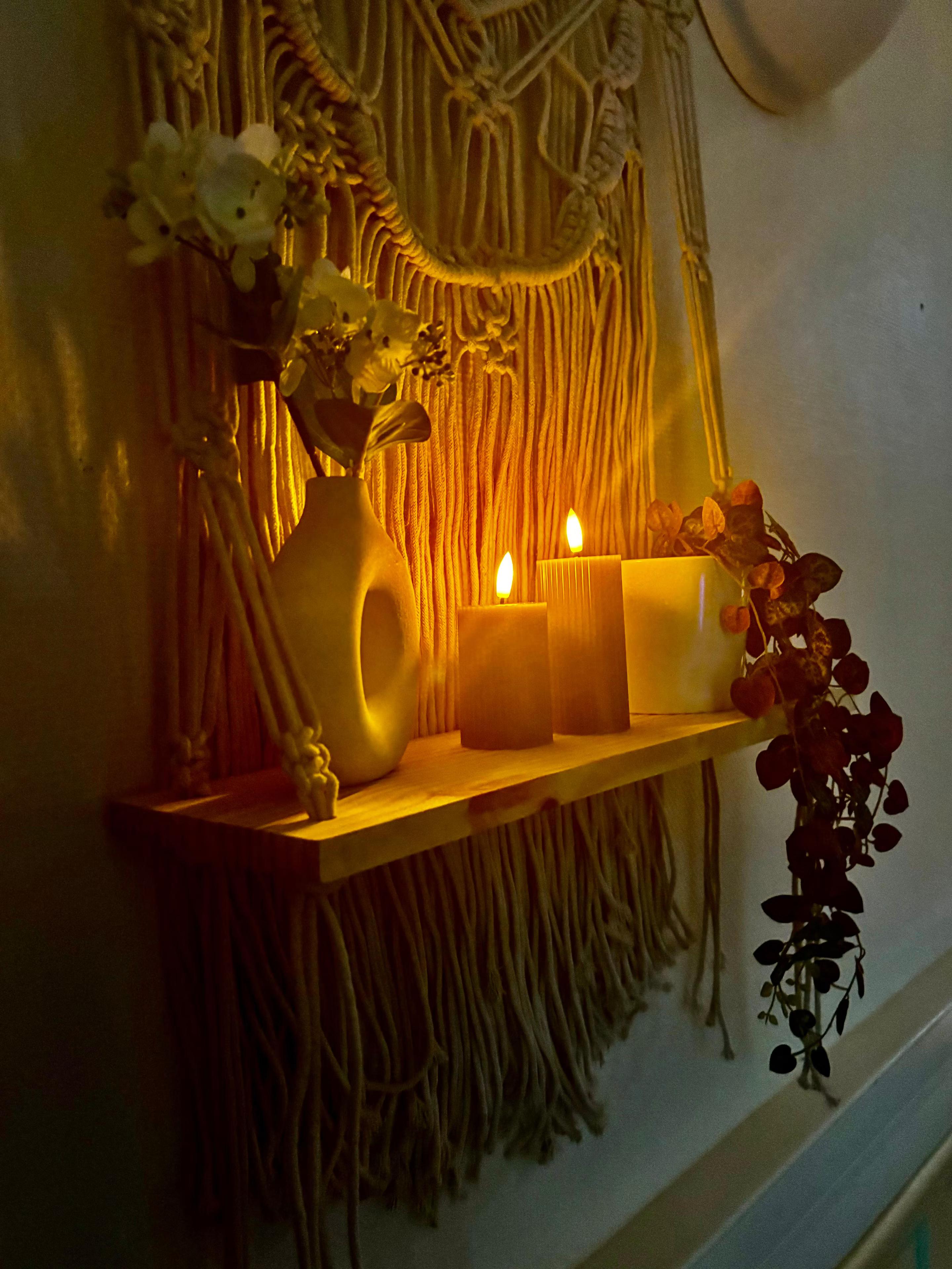Étagère en bois avec bougies allumées, vase fleuri et plante devant un macramé, ambiance tamisée du spa MCH